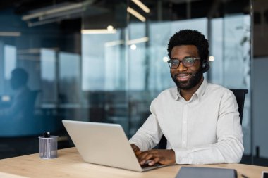 Neşeli bir ofis çalışanı, parlak bir çalışma alanında dizüstü bilgisayar kullanarak kulaklık takıyor. İş, teknoloji, müşteri desteği ve iş yeri görüntüleri için mükemmel..