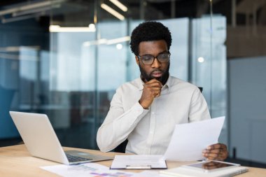 Çağdaş bir ofiste bir işadamı masasında evrakları dikkatle inceler. Bir dizüstü bilgisayar ve akıllı telefon da dahil olmak üzere modern araçlarla çevrelenmiş olup, bir verimlilik ortamını vurguluyor.