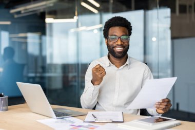 Ofisteki masasında çalışırken özgüven ve başarısını ifade eden profesyonel bir adam. Belgeleri elinde tutuyor ve bir dizüstü bilgisayar kullanıyor. Verimli ve motive edici bir iş tavrı sergiliyor..