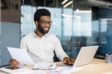 Kendine güvenen bir işadamı evrak işlerini gözden geçirir ve modern ofis ortamında dizüstü bilgisayar kullanır..