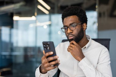 Ofiste akıllı telefonuna dikkatle bakan düşünceli ve odaklanmış bir insan. Profesyonel olarak giyinmiş ve cam panelleri olan çağdaş bir iş yerine yerleştirilmiş.