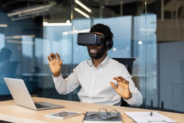 Modern ofis alanındaki gelişmiş VR kulaklıklarıyla sanal gerçekliği deneyimleyen bir iş adamı. Teknoloji, yenilik ve araçlar olarak vurgulanan gelecek kavramları üretkenliği artırır