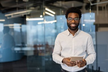 Modern bir ofis ortamında elinde tablet tutarken güven ve dostluk gösteren profesyonel bir adam. Üretken bir çalışma alanında liderlik, teknoloji ve yenilik sunar