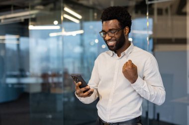 Modern bir ofiste gülümseyen profesyonel bir adam telefonunu tutarken kutlama yapar. İmaj, iş ortamında başarı, ilham ve pozitif enerjiyi temsil eder..