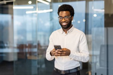 Gözlüklü profesyonel bir adam, cam duvarlı bir ofis ortamında gülümserken bir akıllı telefon tutar ve üretkenlik ve güven anı yakalar..