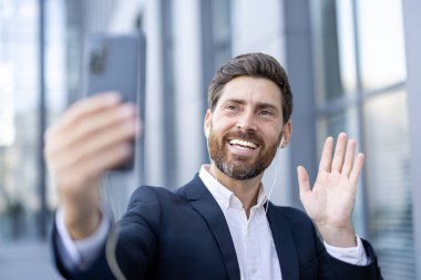 Takım elbiseli neşeli bir adam açık havada bir video görüşmesi yapıyor, el sallıyor ve gülümsüyor, modern iletişimi gösteriyor..