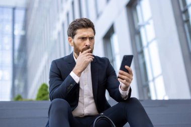 Resmi takım elbiseli sakallı bir iş adamı akıllı telefonundaki bilgileri analiz ederek düşünceli ve endişeli bir tavır sergiliyor..