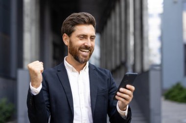 Kendine güvenen bir iş adamı sevinçlidir ve akıllı telefonunu kullanarak başarısını kutlar. Kentsel zemin onun profesyonel ve modern davranışlarını vurguluyor.