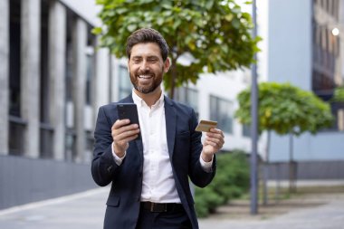 Akıllı telefon ve kredi kartı kullanan, çevrimiçi alışveriş veya dijital işlemleri temsil eden, modern şehir ortamında parlak ve güneşli bir ambiyansla duran, profesyonel kıyafetli bir adam..