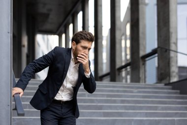 Takım elbiseli bir işadamı endişeli ve iyi görünmüyor. Merdivenlerde tırabzanlara tutunuyor. Hastalık ya da rahatsızlık taşıyor..