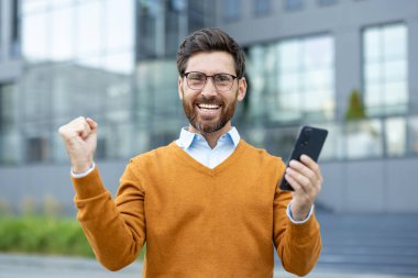 Günlük giysiler içinde mutlu bir adam ofis binalarının yakınında elinde akıllı bir telefon tutarken seviniyor..