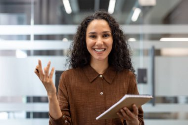 Görüntü, profesyonel bir kadının iş görüşmesi sırasında güven içinde iletişim kurduğunu gösteriyor. Bir defter tutuyor, jestler yapıyor ve içten bir şekilde gülümsüyor. Etkili sunum yetenekleri sergiliyor.