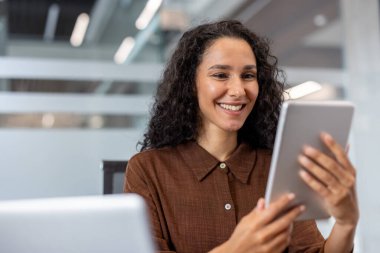 Neşeli bir kadın, parlak, profesyonel bir çalışma alanında tabletle etkileşime girer. Çağdaş ortam ve gülüşü pozitiflik, bağlantı ve verimlilik taşıyor.