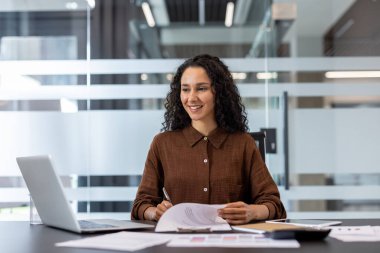 Profesyonel bir birey, belgeleri incelerken bir dizüstü bilgisayar kullanarak masada oturur. Dekor, profesyonel ve çağdaş bir ortam yaratmak için cam duvarları olan modern bir ofis alanı içeriyor..