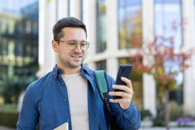 Dışarıda genç bir adam rahat bir ifadeyle akıllı telefonuna bakıyor, sırt çantası ve tablet taşıyor.