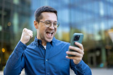 Gözlüklü ve mavi gömlekli neşeli bir adam telefonunu şehir merkezinin arka planında kullanırken zaferini kutluyor. Bu ifade mutluluğu, zaferi ve dijital bağlantıyı yakalar..