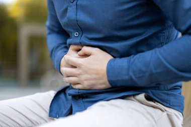 Mavi gömlekli ve bej pantolonlu bir şahıs midesini dışarıda tutuyor. Muhtemel karın ağrısı ya da rahatsızlık belirtisi. Ortam sıradan, ruh hali de sağlık sorunlarıyla ilgili..