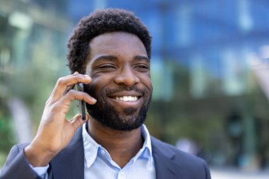 Takım elbiseli bir işadamı dışarıda cep telefonuyla konuşurken gülümsüyor, profesyonel bir ortamda güven ve iletişimi resmediyor..
