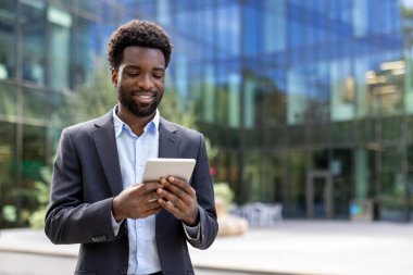 Neşeli, profesyonel bir adam takım elbise içinde elinde bir tablet aygıtla güneşli bir günde şehir merkezindeki modern ofis binasının önünde duruyor. Pozitif bir iş ve teknoloji temasını yansıtıyor..