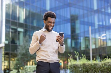 Neşeli bir adam ofis binasının dışındaki akıllı telefonunu kontrol ederken sevinçle tepki veriyor..