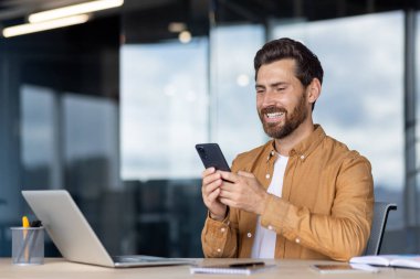 Akıllı telefonu ve parlak çalışma alanında dizüstü bilgisayarı olan neşeli, profesyonel bir çoklu görev. Üretkenliği, modern teknolojiyi ve iş veya uzaktan çalışma için uygun pozitif bir atmosferi yakalamak