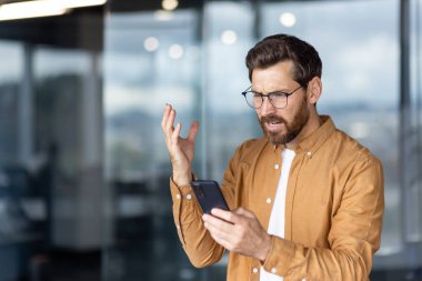 Çağdaş bir ofiste akıllı telefonunu izlerken gözlük takan ve sıradan jestler yapan bir adam. Endişeli ya da hayal kırıklığına uğramış görünüyor, muhtemelen haberlere tepki veriyor.