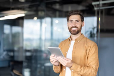 Kendine güvenen bir profesyonel, iyi aydınlatılmış modern bir ofis alanında gülümseyen bir tablet tutar. İş, teknoloji, verimlilik ve profesyonel ve kurumsal liderlik kavramları için mükemmel.
