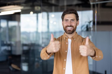 Sakallı, gülümseyen bir adam, parlak ve profesyonel ofis ortamında olumlu ve kendine güvenini gösteriyor..