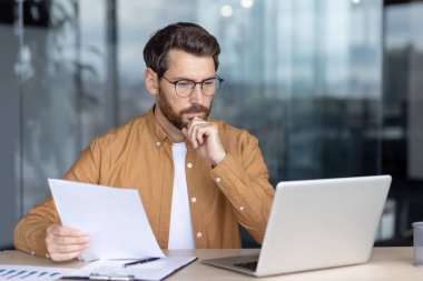 Gözlük takan düşünceli bir adam elinde bir kağıt tutarken elinde dizüstü bilgisayarla, parlak ve modern bir ofis ortamında odaklanmış ve dalgın görünüyor..