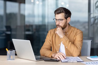 Odaklanmış profesyonel, çağdaş bir ofiste masa başında oturur, görevleri düşünürken bir dizüstü bilgisayar üzerinde çalışır. Atmosfer, konsantrasyon, verimlilik ve kendini işe adamayı iletir..