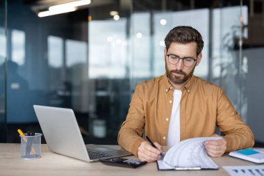 Modern bir ofiste masa başında oturmuş, odaklanma ve kararlılık hissiyle belgeleri dikkatle inceleyen bir profesyonel. Sahne bir dizüstü bilgisayar, hesap makinesi ve yazma gereçleri içeriyor.