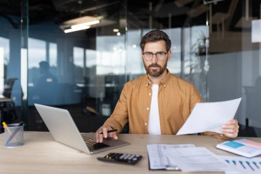 Bir iş adamı dizüstü bilgisayarı ve modern ofis ortamında belgeleri olan bir masada oturuyor. Evrakları odaklanmış bir ifadeyle inceleyip profesyonel ve analitik çalışma tavrı sergiliyor..
