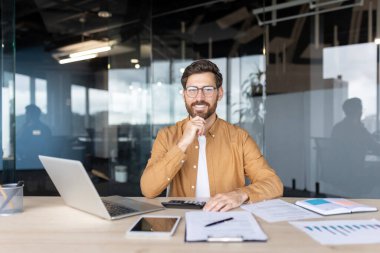 Parlak bir ofisin içinde, masasında oturan gülümseyen bir adam..