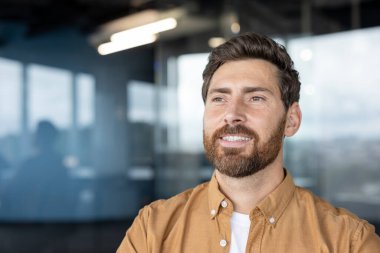 Büyük pencereli, parlak, çağdaş bir ofis alanında sıradan bir tişört giyen gülümseyen adam. Profesyonel ortam ve onun olumlu ifadesi güven ve başarıyı yansıtıyor.