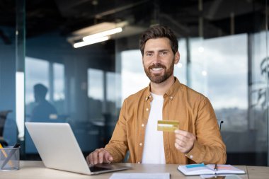 Modern ofis ortamında gülümseyen bir adam dizüstü bilgisayarını kullanırken elinde kredi kartı tutuyor. Görüntü, profesyonel bir çalışma alanında finans, ticari işlemler ve çevrimiçi alışverişi temsil etmektedir..