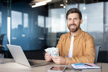 Neşeli bir adam elinde dizüstü bilgisayarla bir masada otururken ve gerekli işleri organize ederken, profesyonel ofis alanındaki mali başarıyı ve verimliliği simgeleyen banknotlar tutuyor..