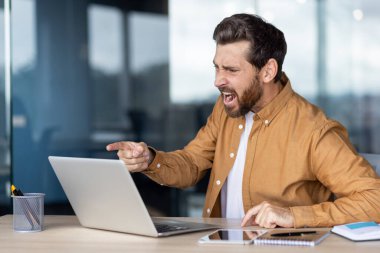 Modern ofis ortamında dizüstü bilgisayara güçlü bir şekilde el kol hareketi yapan kendine güvenen bir iş adamı. Yoğun tepkisi hayal kırıklığını ya da kararlılığı gösteriyor.