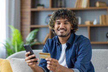 Çevrimiçi işlemler için kredi kartı ve akıllı telefonunu kullanırken modern oturma odasında oturan neşeli bir adam. Ortam, rahat bir atmosfer ve dijital ticarete olumlu bir yaklaşım izlenimi veriyor..