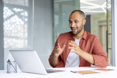 Kırmızı tişörtlü gülümseyen adam, iyi aydınlatılmış modern ofis ortamında dizüstü bilgisayar kullanarak konuşma sırasında coşkuyla el kol hareketi yapıyor..