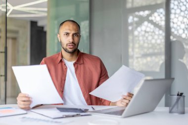 Düşünceli ve odaklanmış bir birey şık bir ofiste dokümanlar ve dizüstü bilgisayarla çalışır. Bu senaryoda modern çalışma alanı, üretkenlik ve profesyonellik temsil edilmektedir..