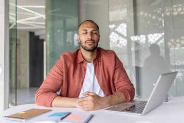 Huzurlu bir birey, bir ofis masasında, gözleri kapalı, rahatlama ve farkındalığı kucaklayarak huzur içinde oturur. Ayarlar, profesyonel bir ortamda sükunet olduğunu gösteriyor.