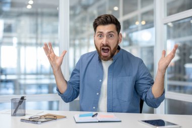 Çağdaş bir ofiste masasında çalışan bir adam şaşkınlığını ya da şaşkınlığını ifade eder. Çalışma alanı, masanın üzerinde notlar ve gözlüklerle modern ve şeffaf bir tasarıma sahiptir..