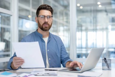 Bir iş adamı elinde belge tutarken bir dizüstü bilgisayar üzerinde çalışıyor, modern cam duvarlı arka planı olan bir ofis masasında oturuyor..