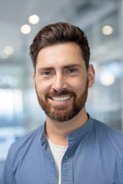 Neşeli bir adam parlak, modern ofis ortamında sıcak bir şekilde gülümser. Doğal ışık, karşılama ve profesyonel atmosferi, pozitifliği ve ulaşılabilirliği artırır..