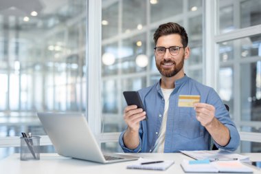 Akıllı telefon ve kredi kartı tutan bir iş adamı. Parlak bir ofis masasında çalışıyor..