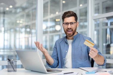 Ofis masasında kredi kartı tutarken hayal kırıklığı gösteren bir adam, endişelerini dile getiriyor..