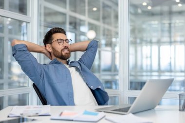 Masasında açık ve modern bir ofis ortamında oturmuş dinlenen bir adam. Yoğun bir çalışma gününde rahatlık ve memnuniyeti yansıtan bir dinlenme anından hoşlanıyor gibi görünüyor..