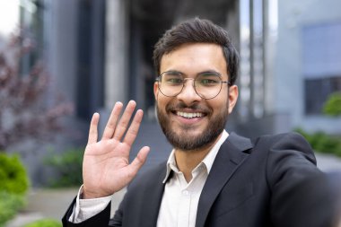Açık havada kameraya el sallarken gülümseyen resmi giyinmiş kendinden emin genç bir adam. İletişim, iş ve sosyal etkileşim kavramları için ideal.