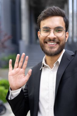 Takım elbiseli, gözlüklü, gülümseyen ve dalgalanan kendine güvenen bir işadamı, kentsel ve profesyonel bir ortamda misafirperver ve cana yakın bir tavır sergiliyor..