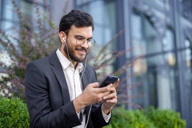 Takım elbiseli iyi giyimli profesyonel bir adam akıllı telefonu ve kulaklıklarıyla dışarıda takılıyor..